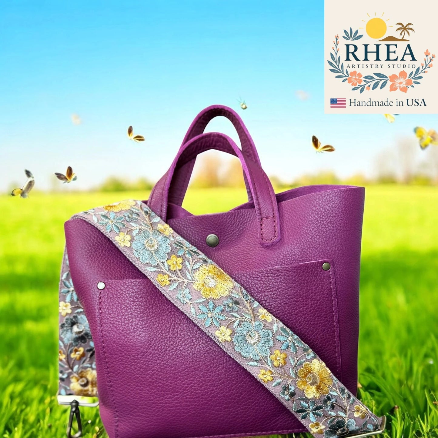 Purple handbag with a floral bag strap on a grassy field background, featuring the RHEA Artistry Studio logo.