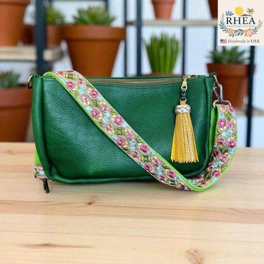 Green handbag with a decorative strap featuring flowers and a yellow tassel on a wooden surface, with 'RHEEA' branding.