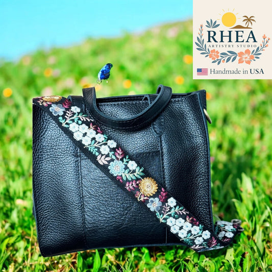 Black leather handbag with floral strap on grass, RHEA Artistry Studio logo visible