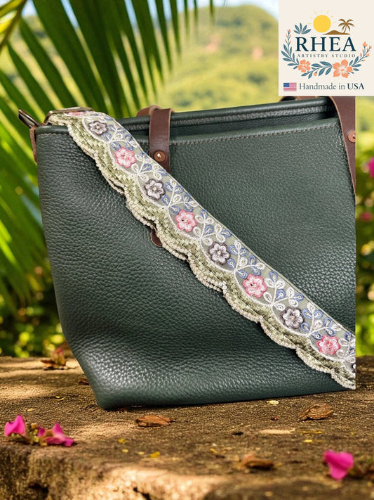 Green handbag with floral lace strap on a wooden surface with a blurred natural background, featuring the RHEA brand.