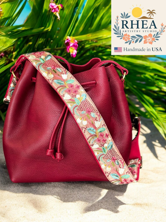 Red leather bucket bag with a floral purse strap on a beach background, featuring the RHEA brand logo.