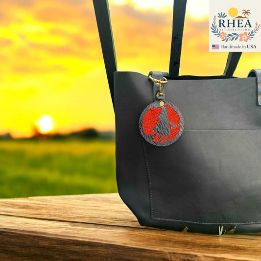 Black bag with a red and gray circular witch leather bag charm against a sunset background, featuring the RHEA brand.
