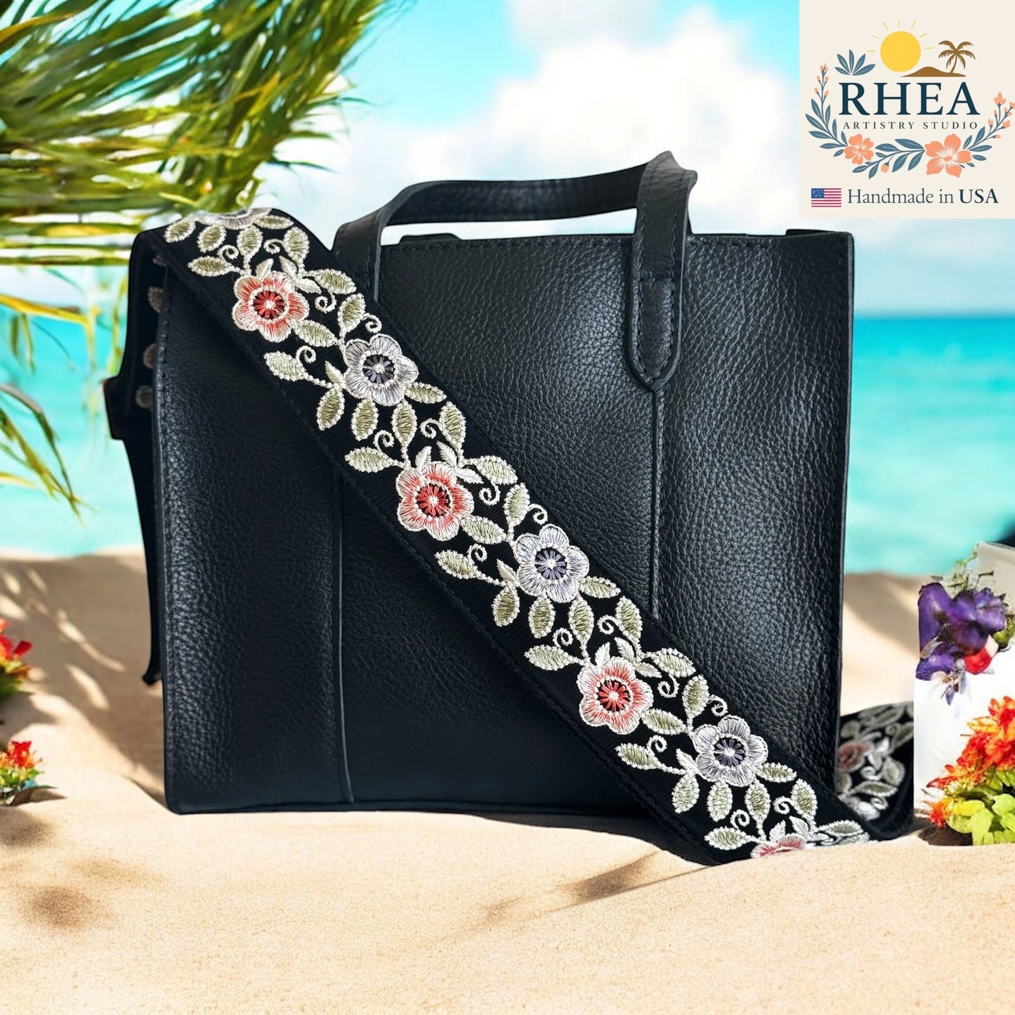 Black handbag with floral purse strap on a beach, RHEA brand visible
