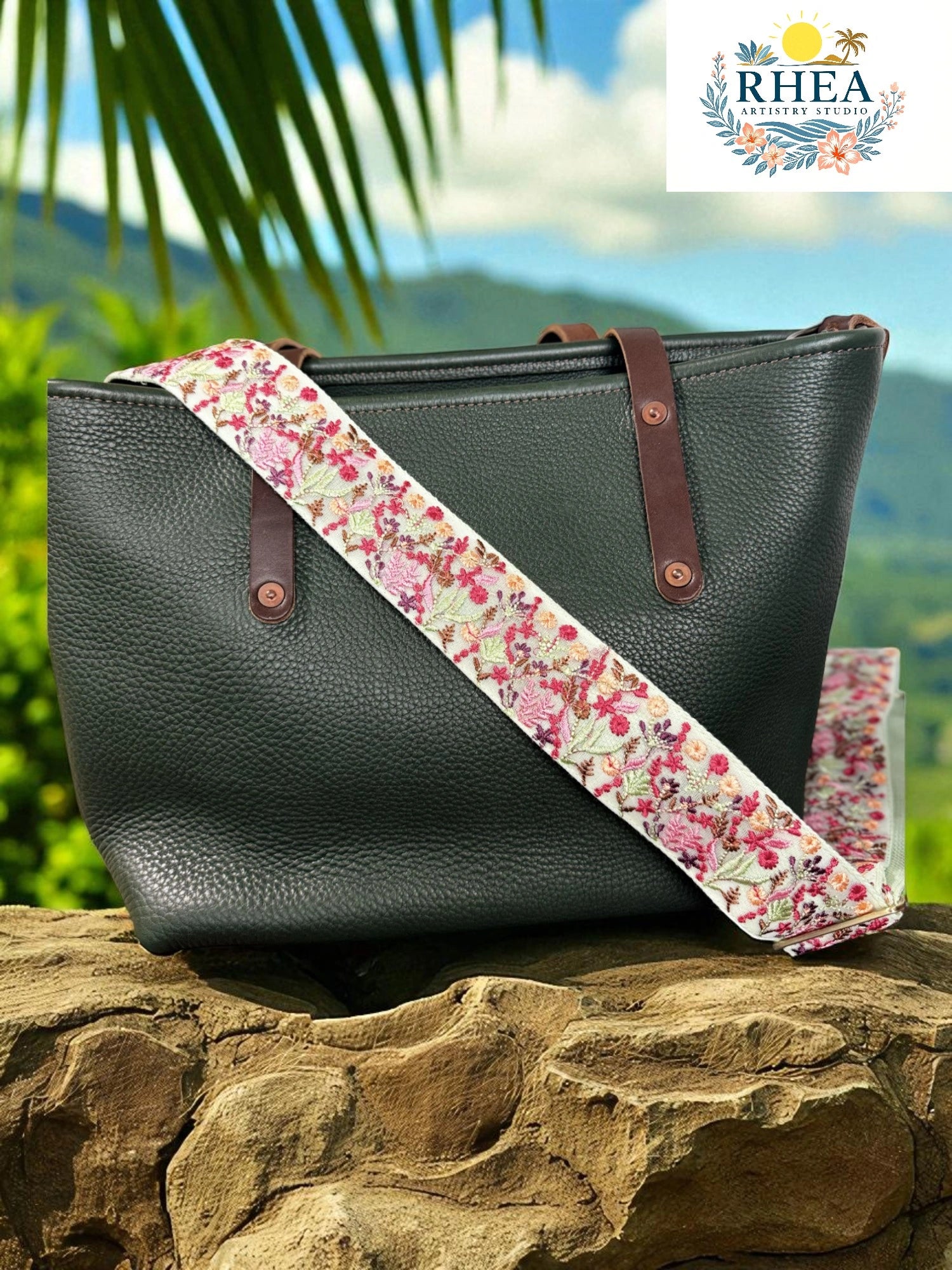 Black leather handbag with a floral strap on a rock with a scenic background, featuring the RHEA brand.