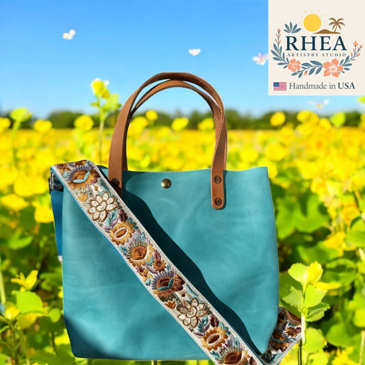 Blue tote bag with floral purse strap in a field of yellow flowers, featuring Rhea Artistry Studio logo.