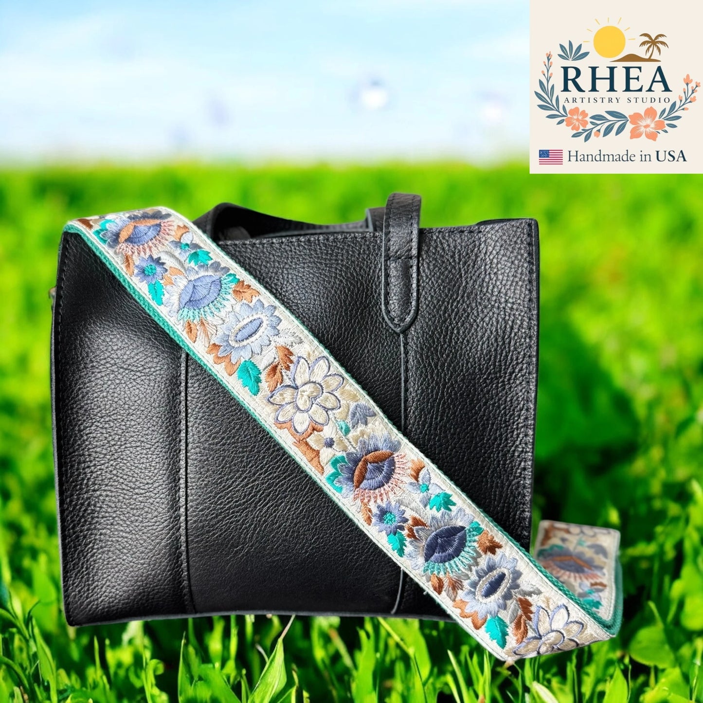 Black leather bag with an embroidered purse strap on a grassy background, featuring the RHEA Artistry Studios logo.