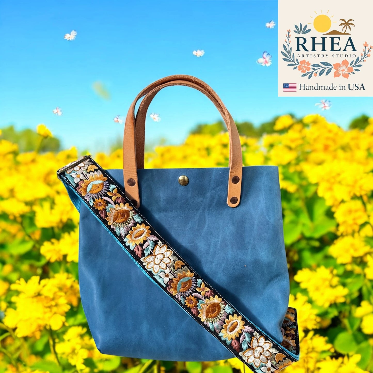 Blue tote bag with floral purse strap against a yellow flower field background, featuring the RHEA Artistry Studio logo.