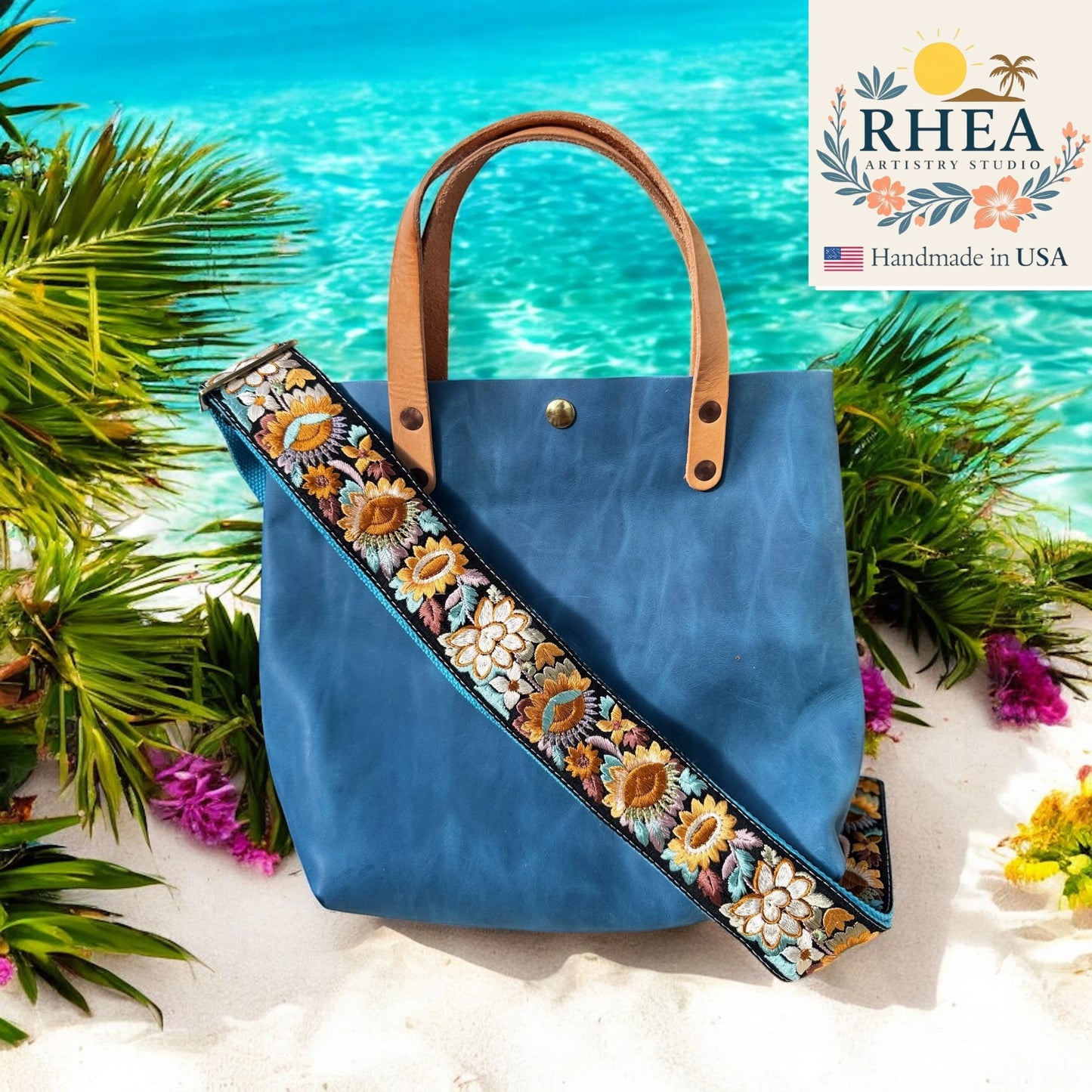 Blue handbag with floral purse strap on a beach background, featuring the RHEA Artistry Studio logo.