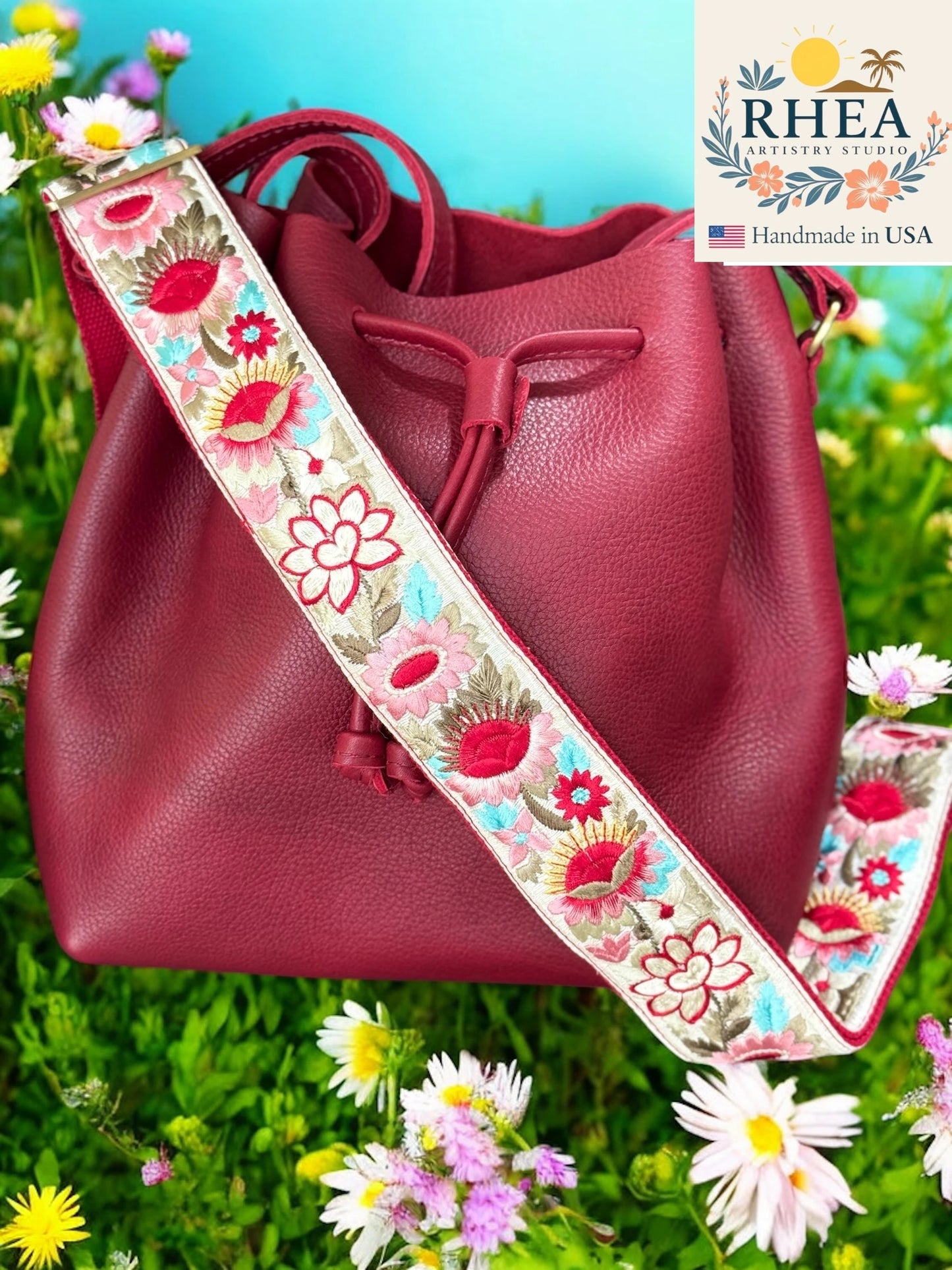 Red handbag with a floral purse strap on a grassy background, featuring the RHEA brand.