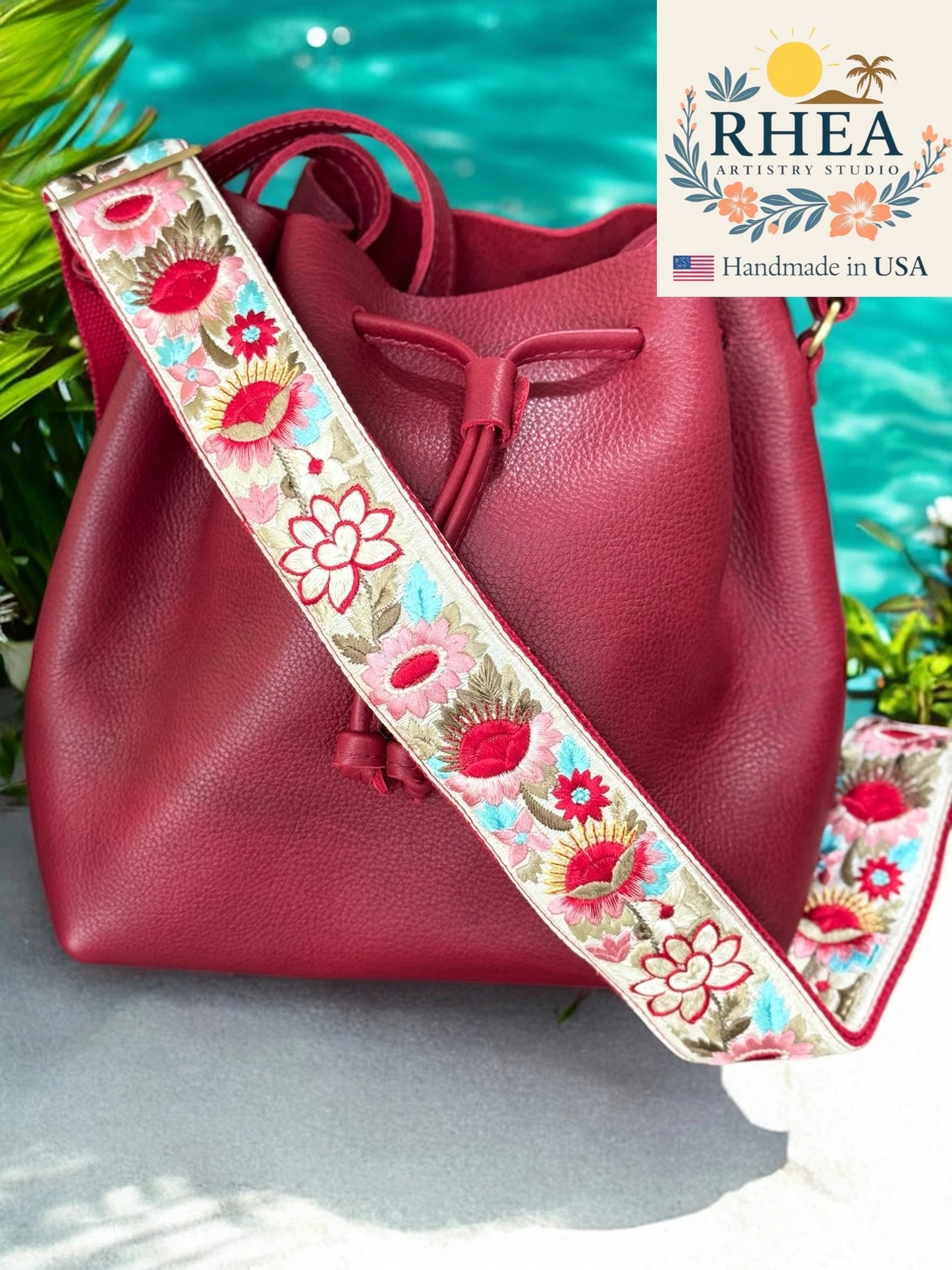 Red handbag with a floral purse strap against a blurred natural background, featuring the RHEA brand.