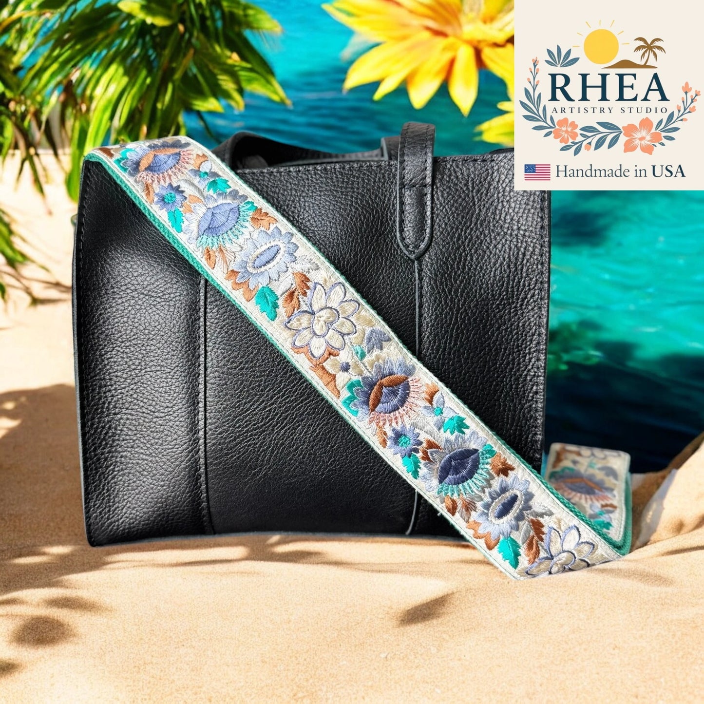 Black handbag with a floral purse strap on sand, flowers, and RHEA Artistry Studio logo.