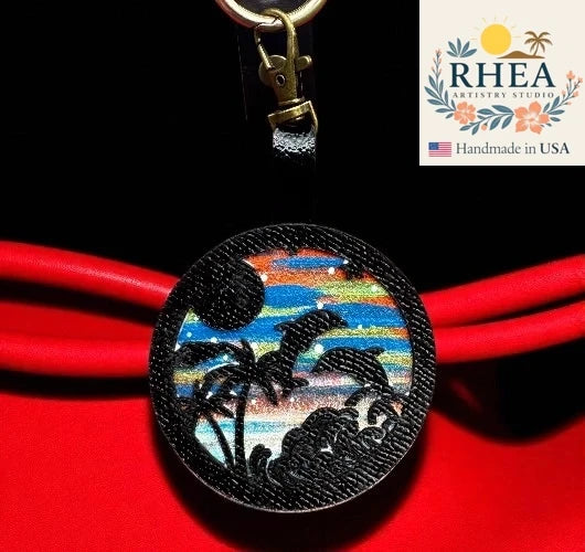 Decorative leather purse charm with sunset design on a red background, featuring the RHEA Artisan Studio logo.
