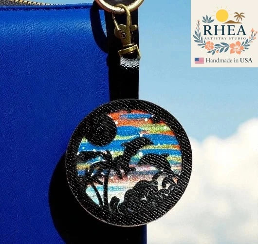 Round leather purse charm with sunset design featuring birds and palm trees, attached to a blue bag.