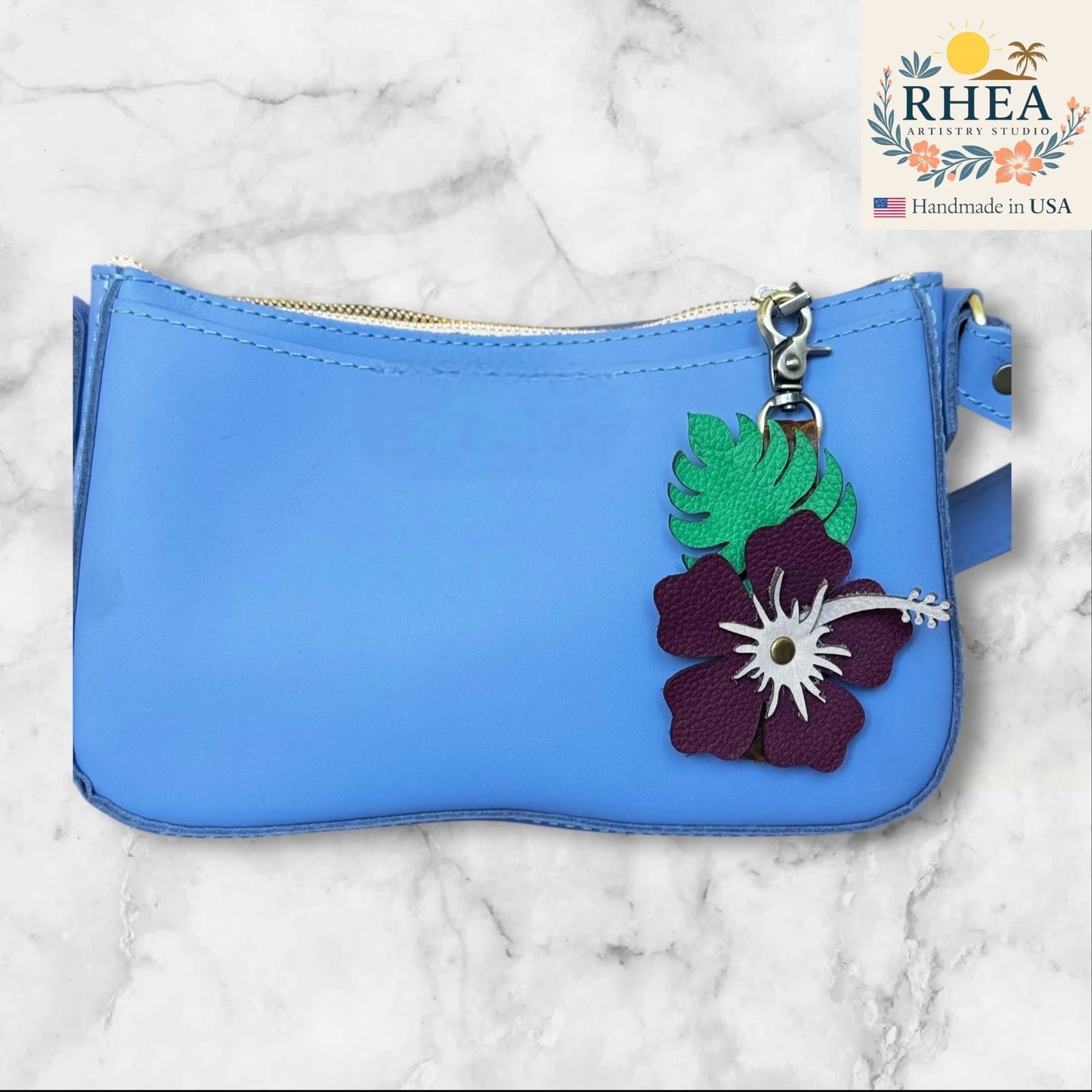 Blue pouch with floral leather purse charm on a marble background, featuring the RHEA brand.