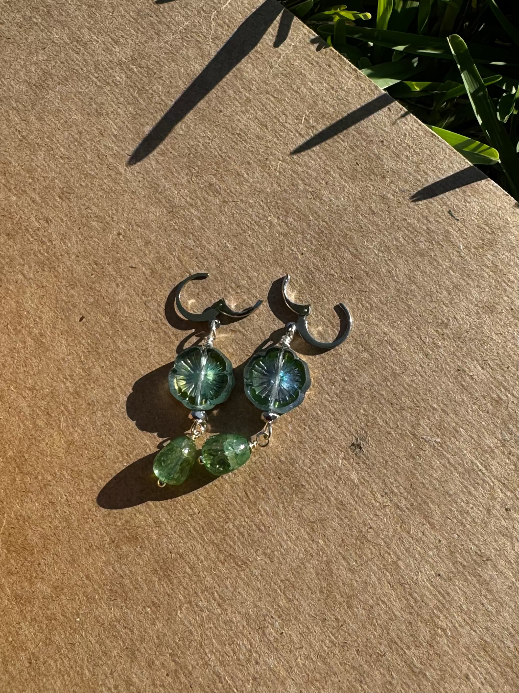 A pair of silver earrings with green gemstone drops and metallic accents placed on a textured surface.