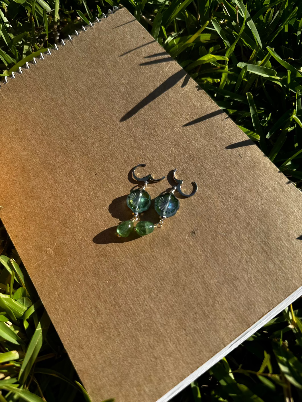 A pair of silver earrings with green gemstone drops and metallic accents placed on a textured surface.