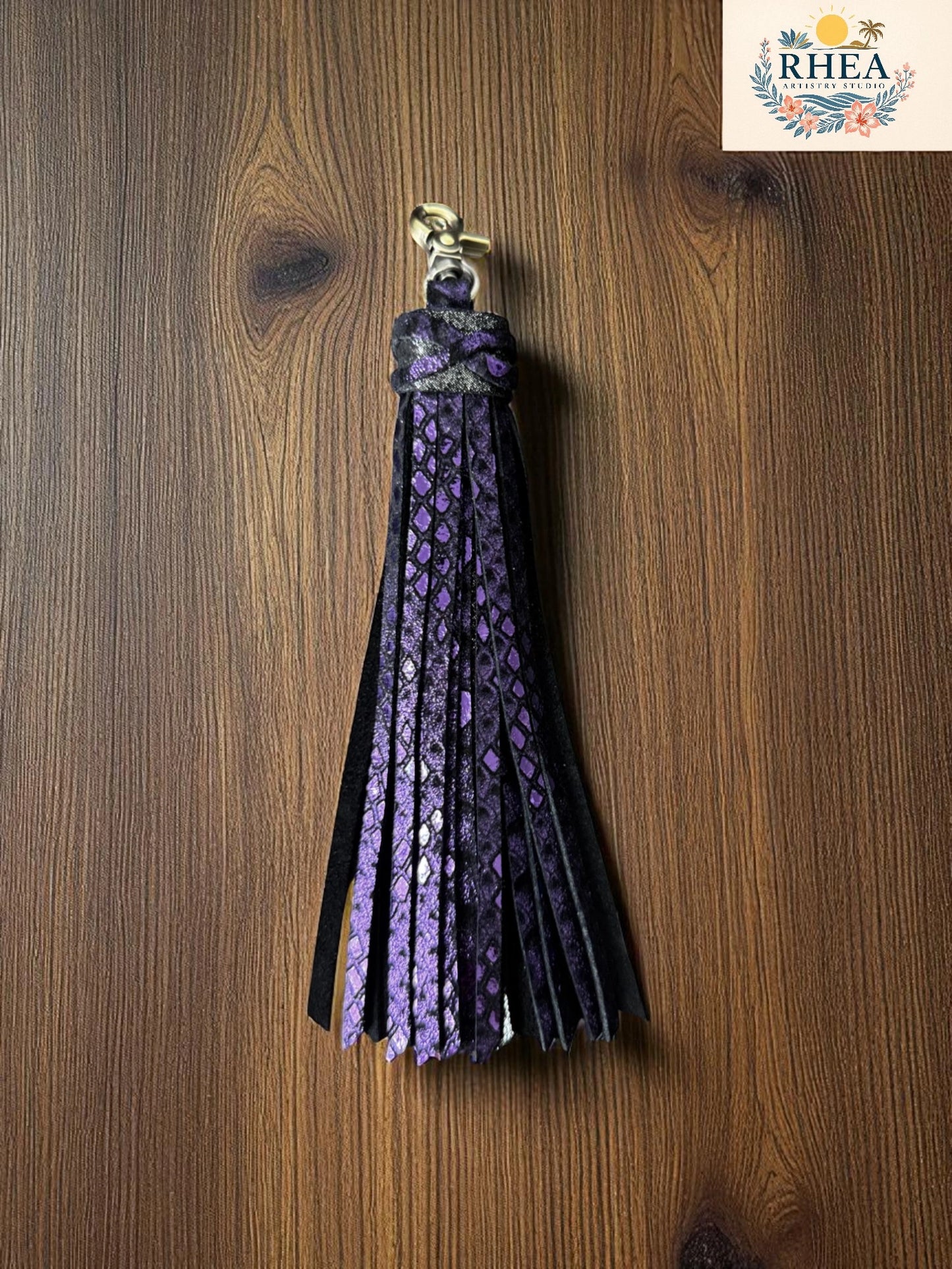 Purple tassel keychain on a wooden surface with RHEA logo in the corner.