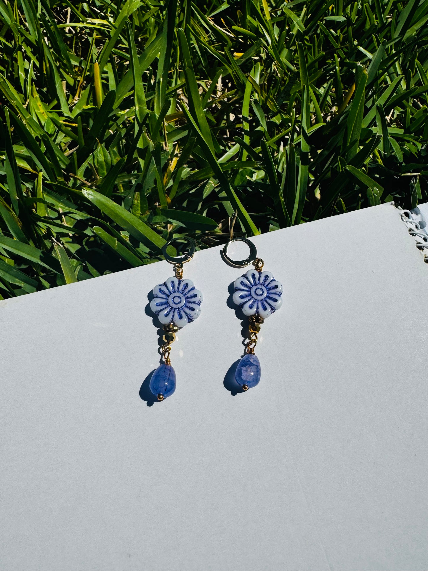 A pair of blue glass drop earrings with gold plated brass findings, displayed on a flat surface with grass and a clear sky in the background.
