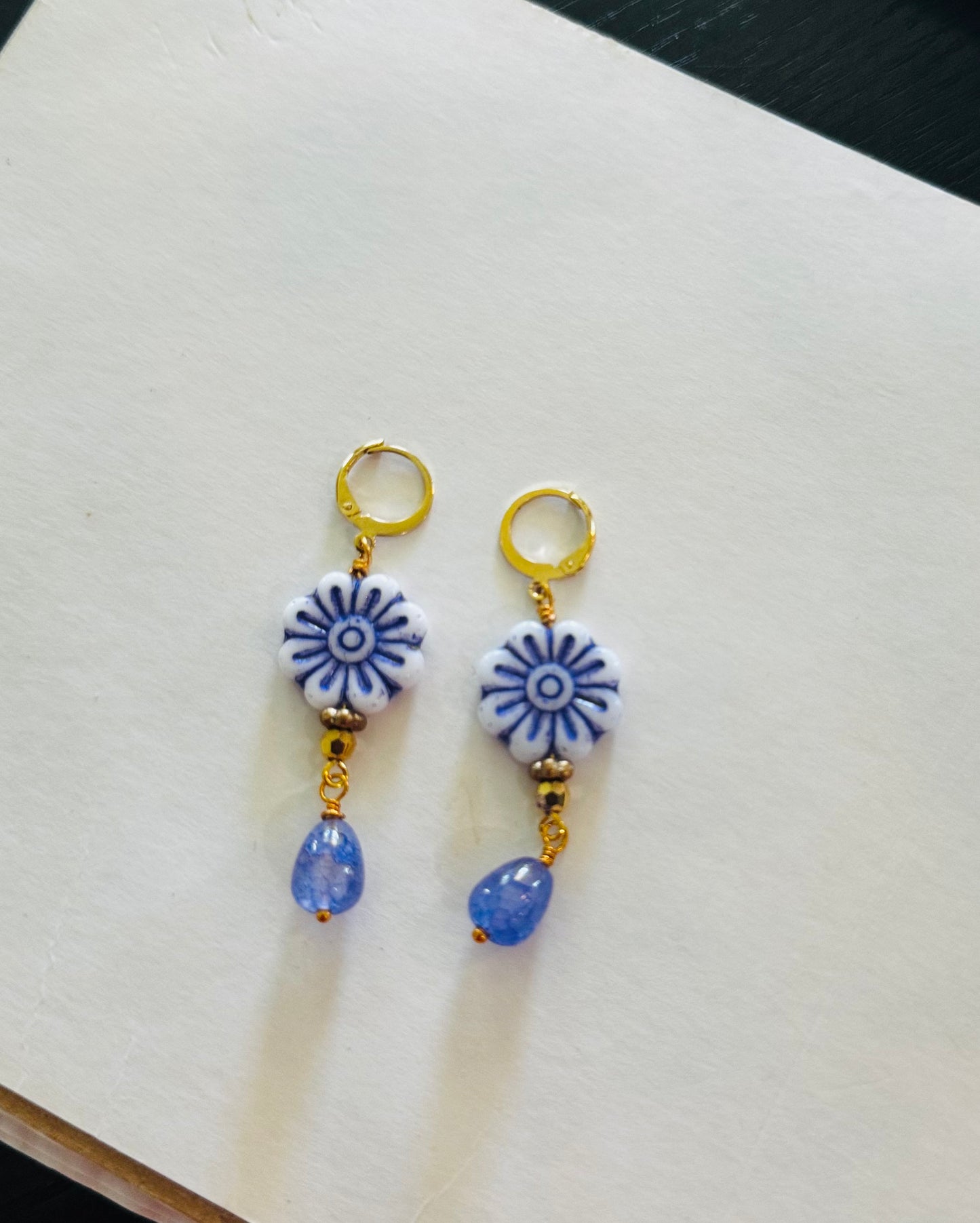 A pair of blue glass drop earrings with gold plated brass findings, displayed on a flat surface with grass and a clear sky in the background.