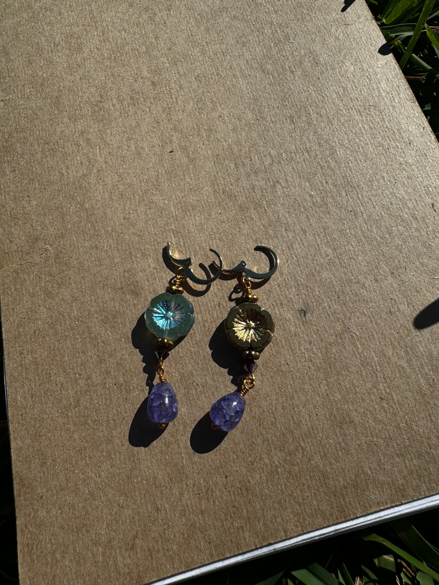 A pair of hibiscus violet drop earrings with gemstones and crystal embellishments, placed on a brown surface.