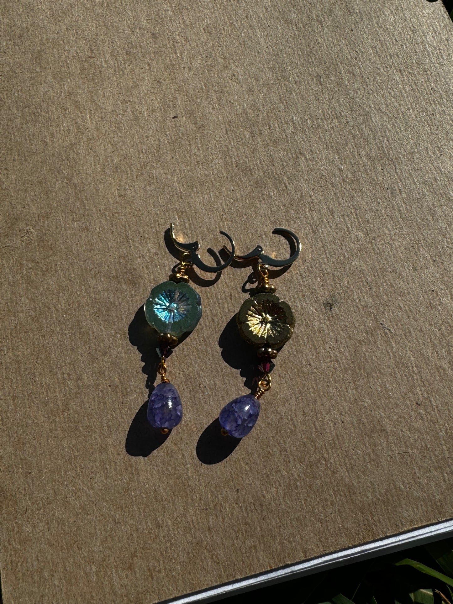 A pair of hibiscus violet drop earrings with gemstones and crystal embellishments, placed on a brown surface.