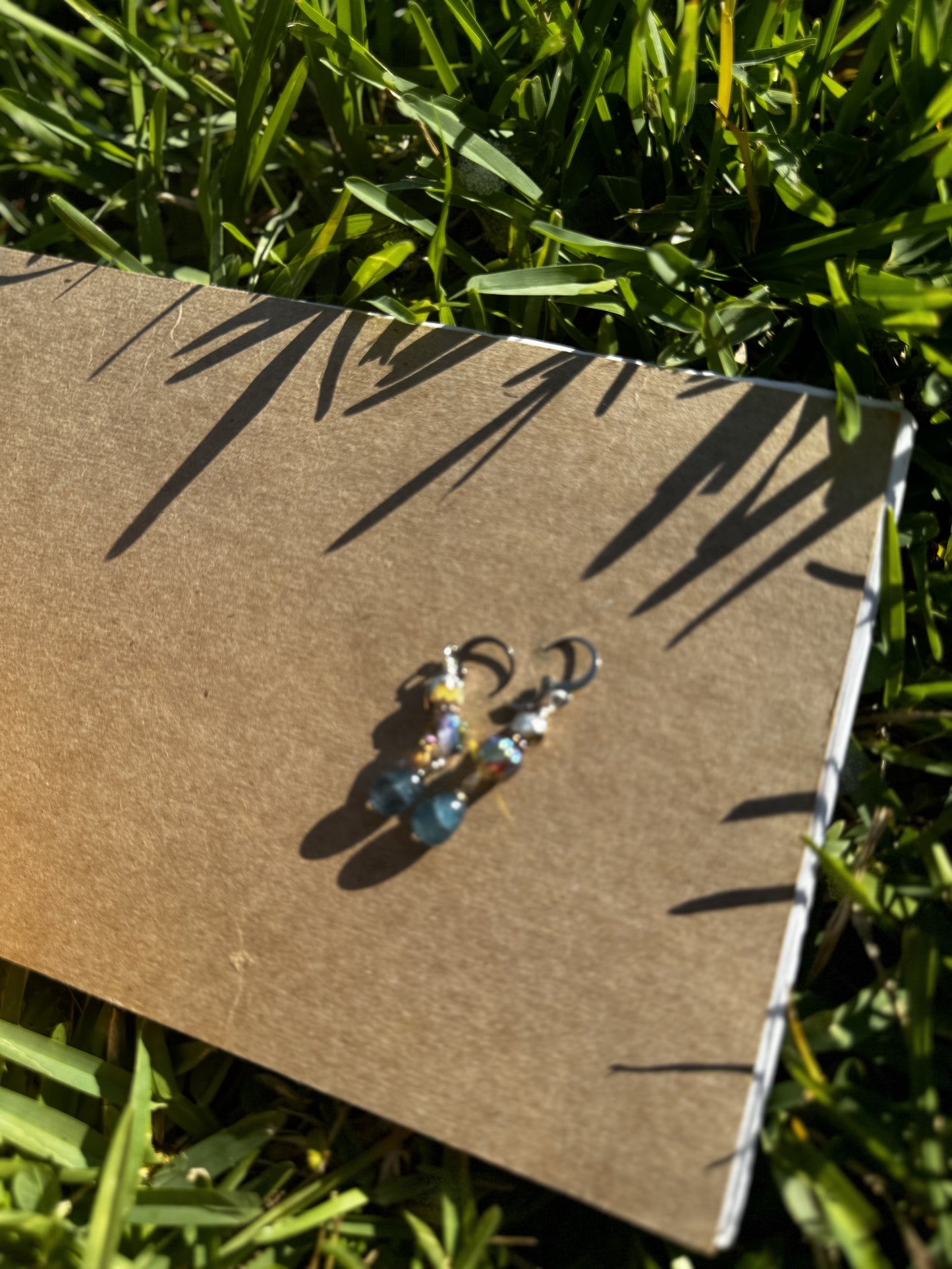 A pair of silver drop earrings with blue and multicolored gemstones, displayed on a brown card placed on grass.