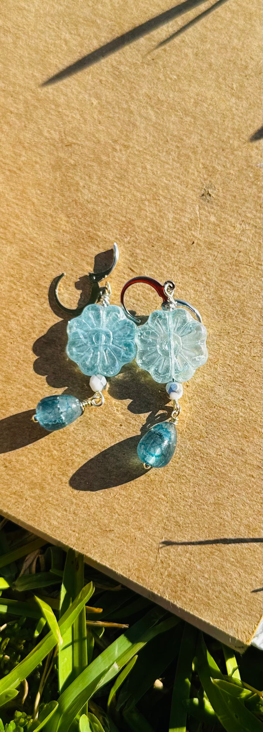 A pair of Daisy Blue Silver Drop Earrings with a blue gemstone, displayed on a brown cardboard surface outdoors.