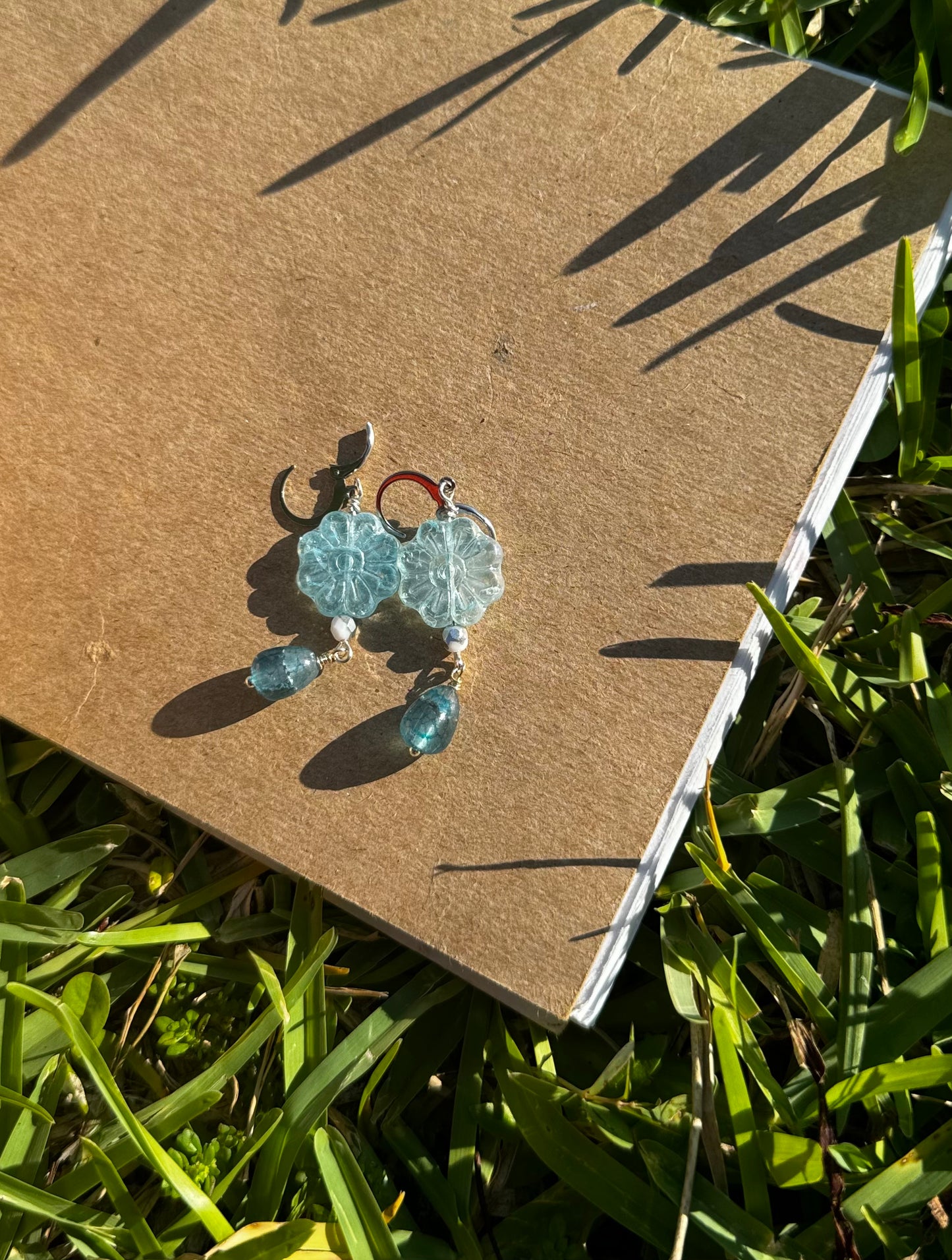 A pair of Daisy Blue Silver Drop Earrings with a blue gemstone, displayed on a brown cardboard surface outdoors.