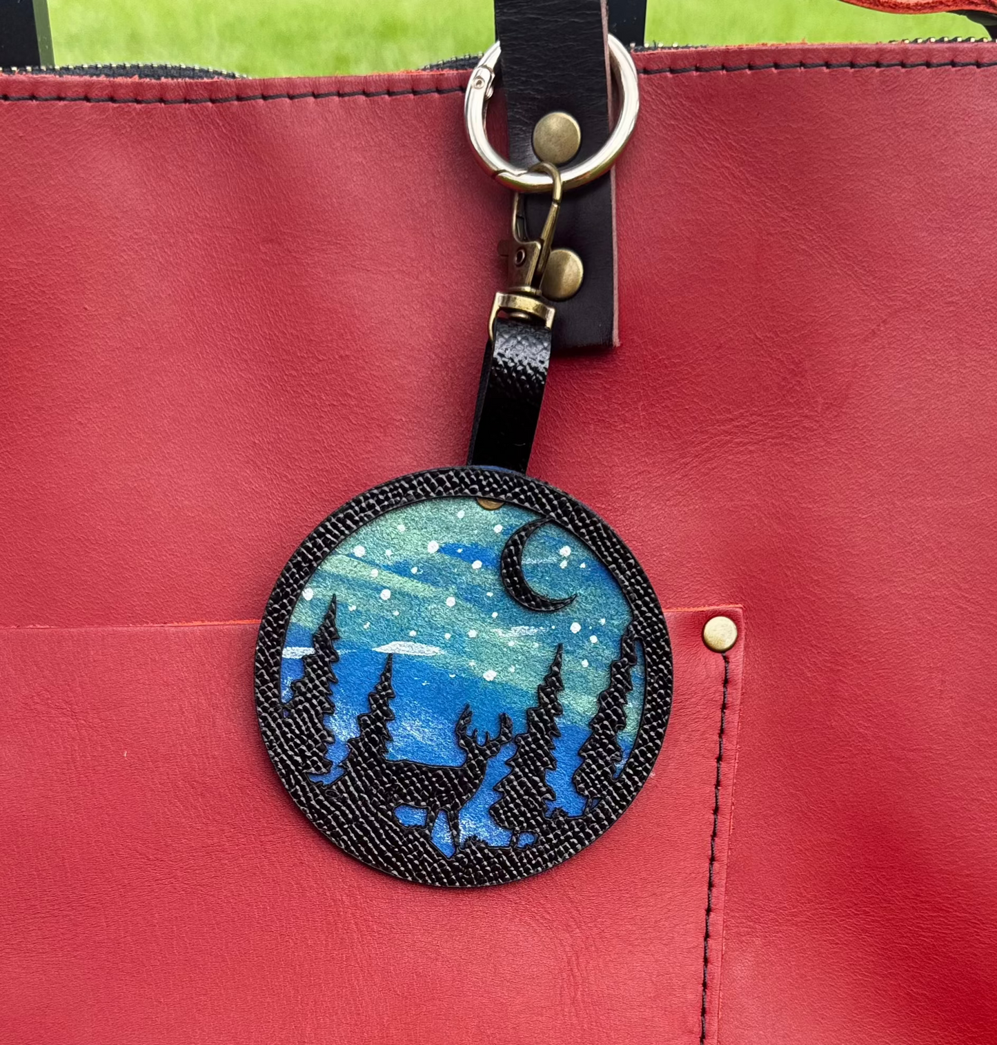 A handpainted leather charm with a starry night landscape, a deer, and a forest design, attached to a red leather bag.
