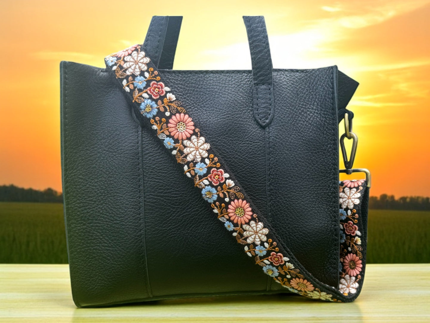 An adjustable strap with daisy flowers embroidery attached to a black bag, with a field and sunset in the background.