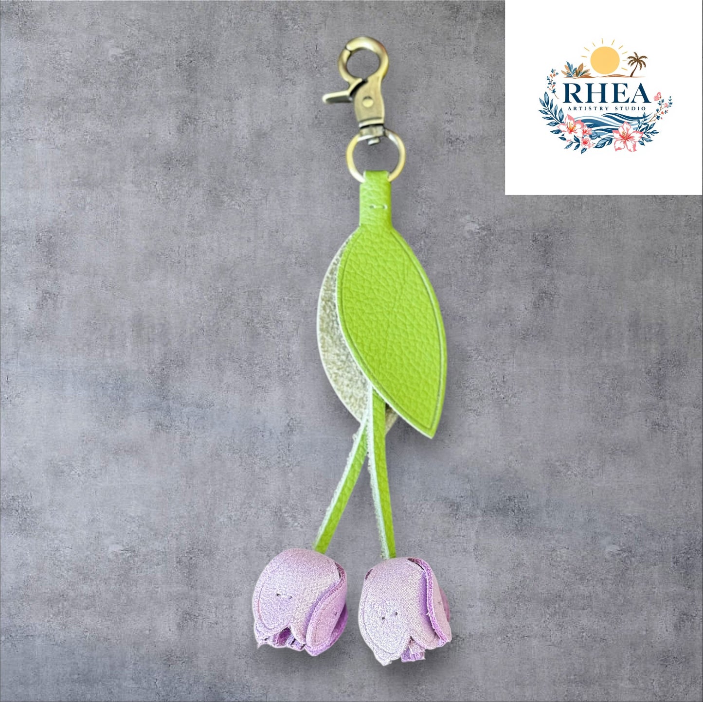 A handmade leather tulip charm with a green leaf and brown floral pattern, attached to a gold lobster clasp on a white marble surface.