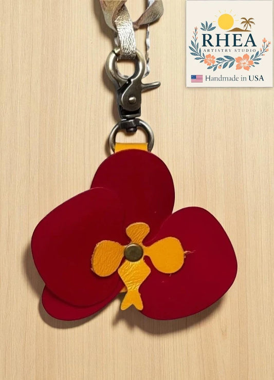 Red and yellow flower-shaped charm with RHEA Artistry Studio branding on a wooden background