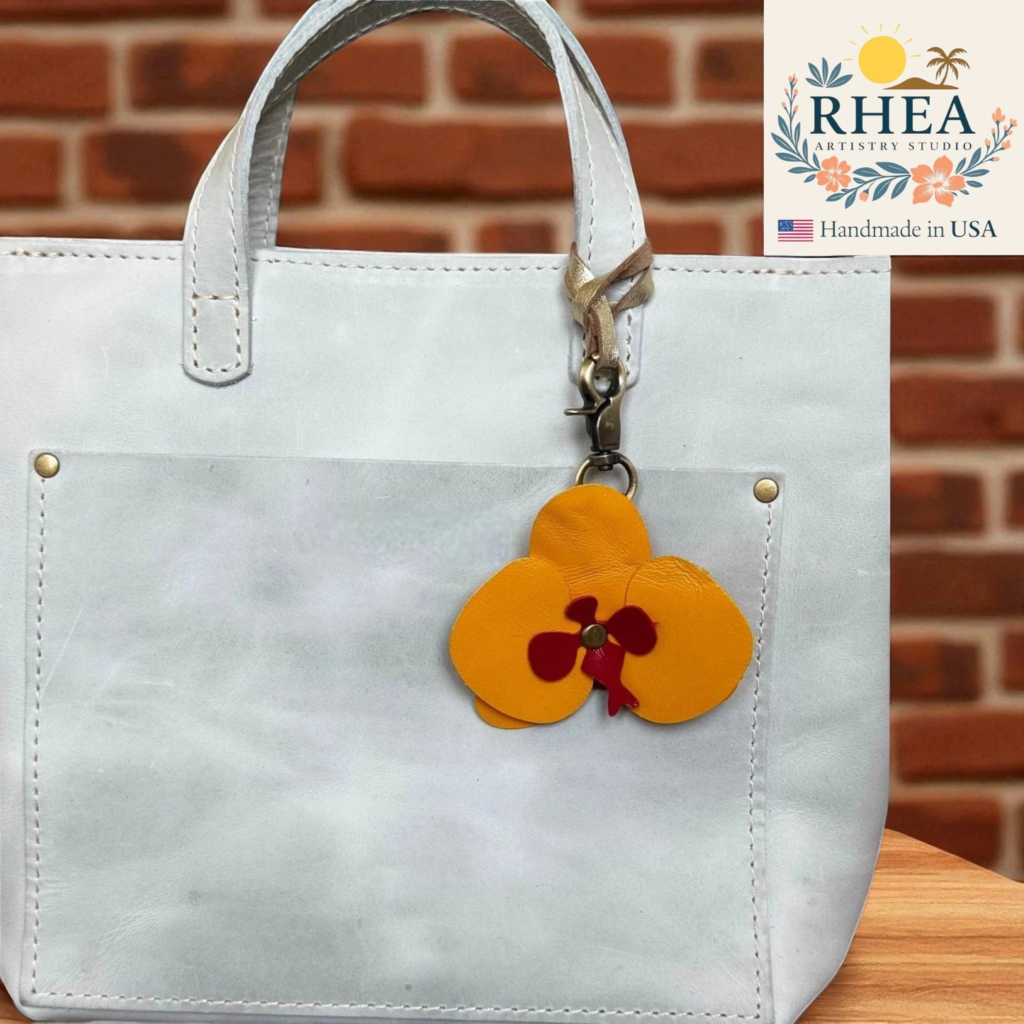 White handbag with a yellow flower charm on a wooden surface and brick wall background, featuring the RHEA Artistry Studios logo.