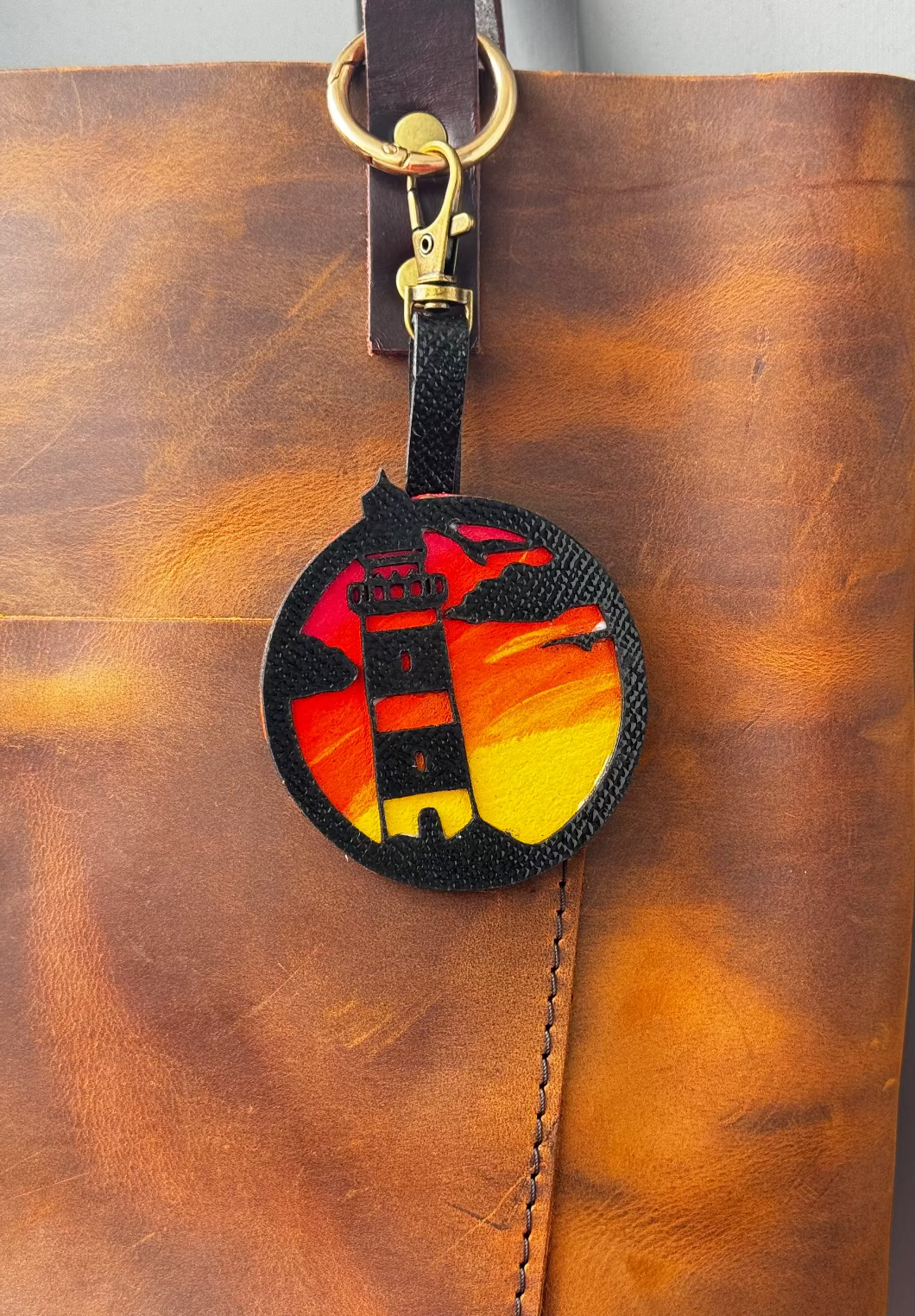 A hand-painted leather charm with a lighthouse design, attached to a brown leather bag with a brass clasp.