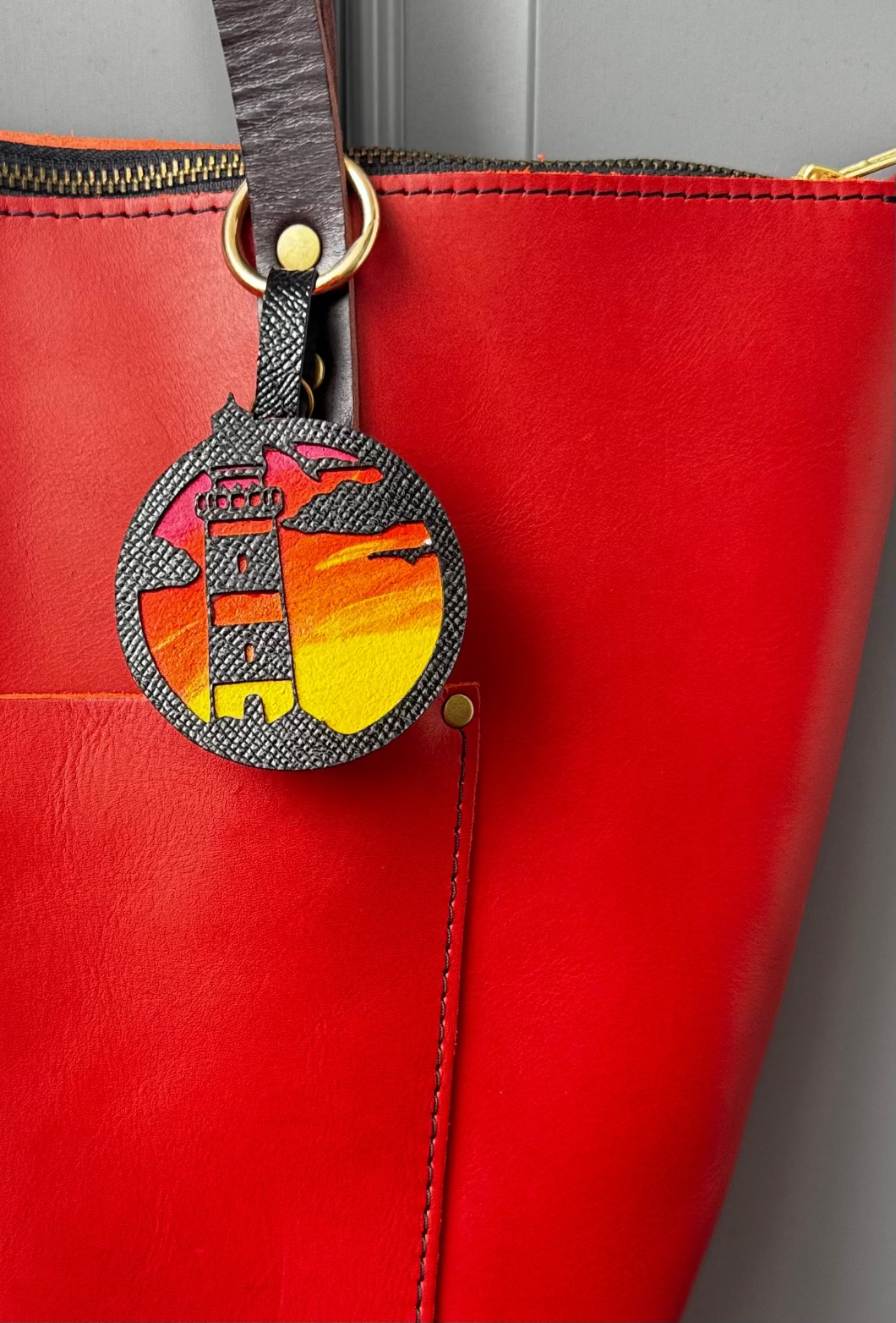 A hand-painted leather charm with a lighthouse design, attached to a red leather bag with a brass clasp.