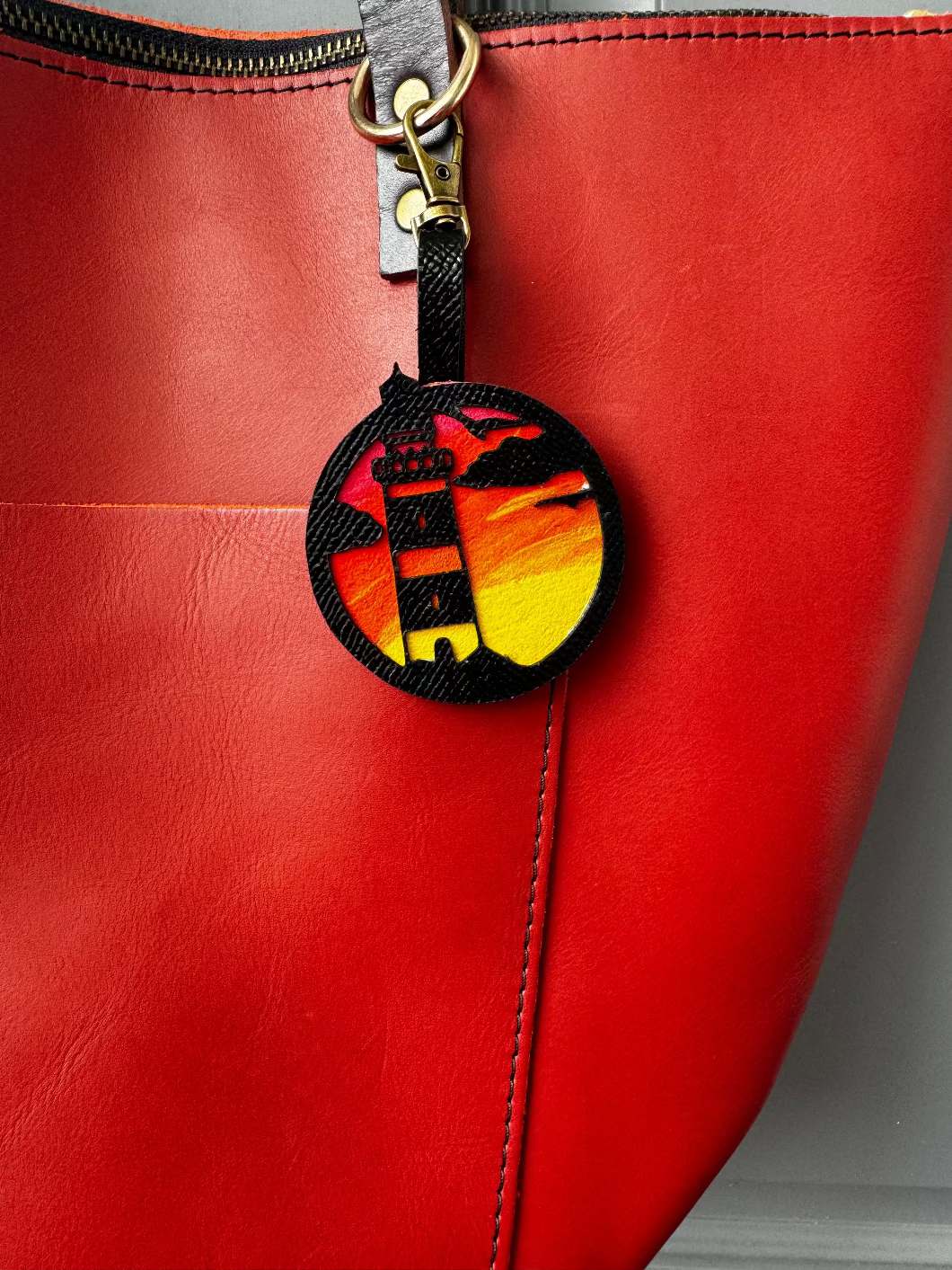 A hand-painted leather charm with a lighthouse design, attached to a red leather bag with a brass clasp.