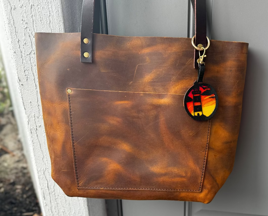 A hand-painted leather charm with a lighthouse design, attached to a brown leather bag with a brass clasp.