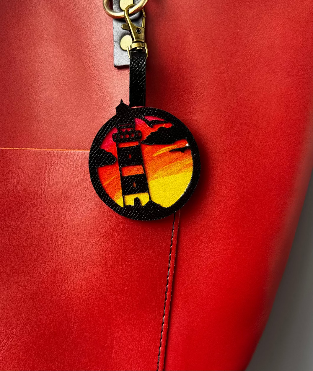 A hand-painted leather charm with a lighthouse design, attached to a red leather bag with a brass clasp.