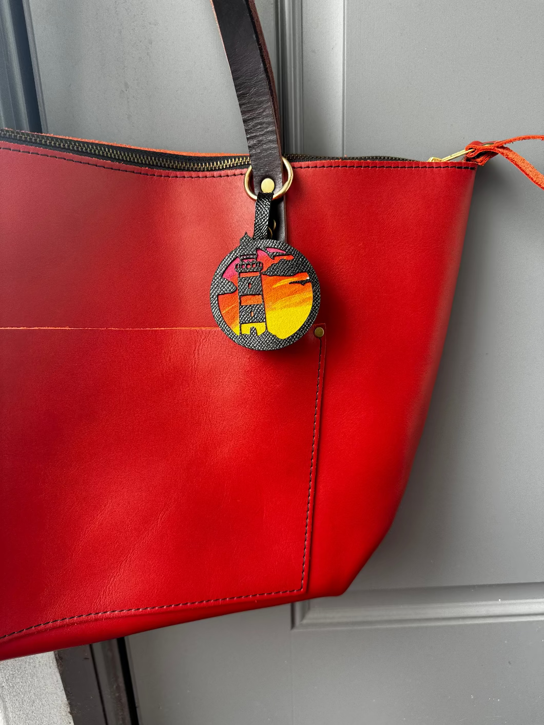 A hand-painted leather charm with a lighthouse design, attached to a red leather bag with a brass clasp.