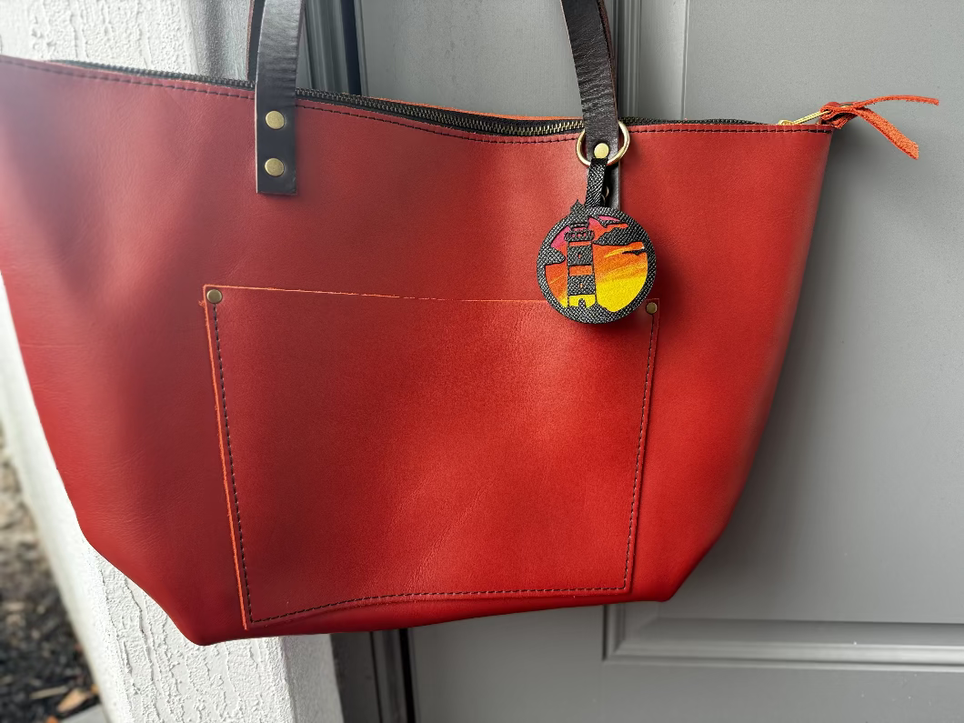 A hand-painted leather charm with a lighthouse design, attached to a red leather bag with a brass clasp.