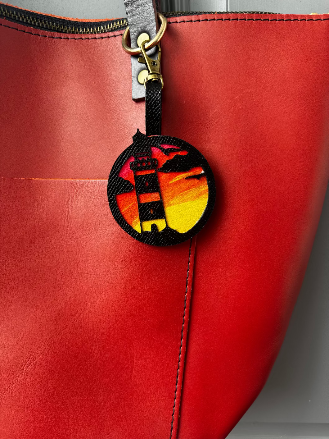 A hand-painted leather charm with a lighthouse design, attached to a red leather bag with a brass clasp.