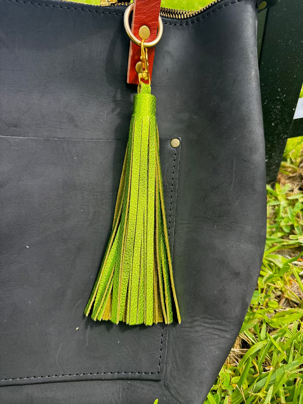 A jumbo leather tassel accessory in fern green color, displayed on a black handbag with a red strap.