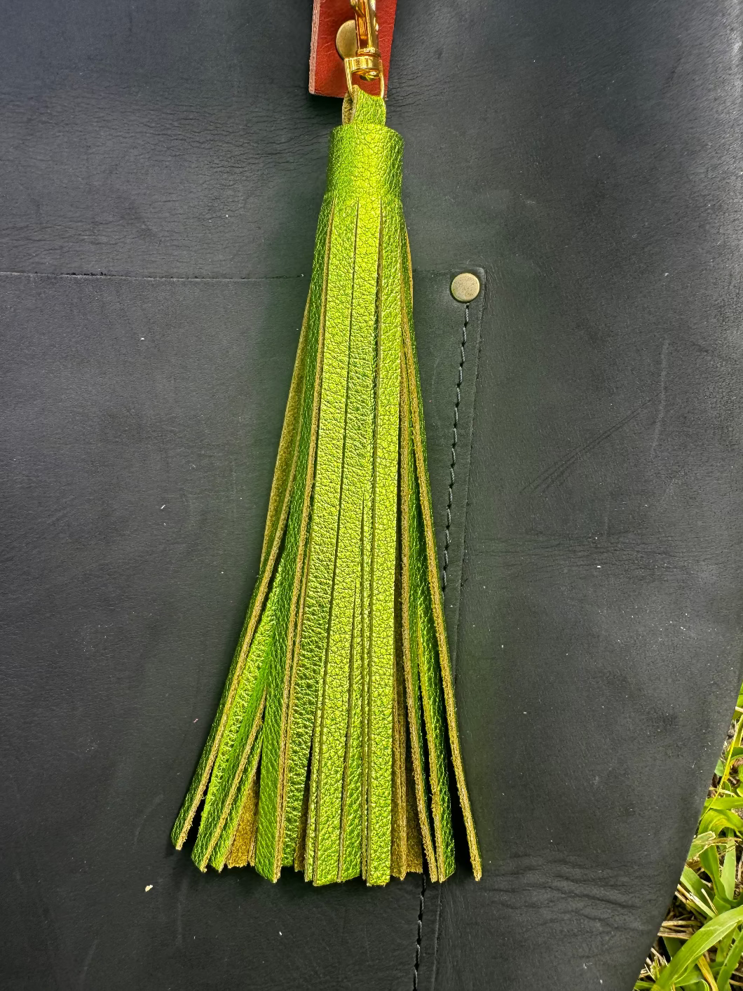 A jumbo leather tassel accessory in fern green color, displayed on a black handbag with a red strap.