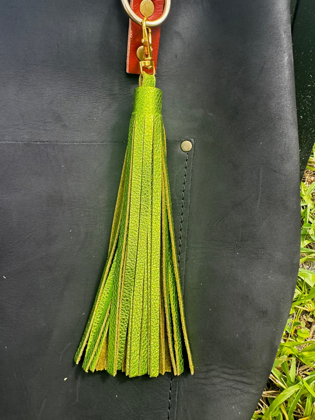 A jumbo leather tassel accessory in fern green color, displayed on a black handbag with a red strap.