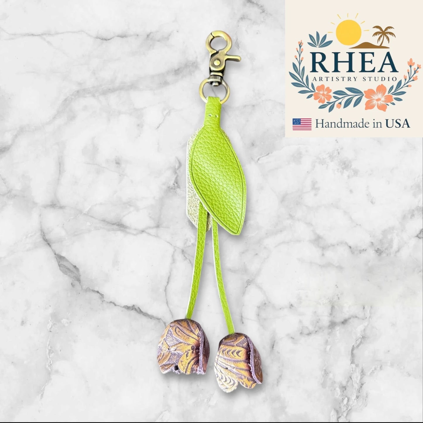 Green leather purse charm with decorative elements on a marble background, featuring the RHEA Artistry Studio logo.