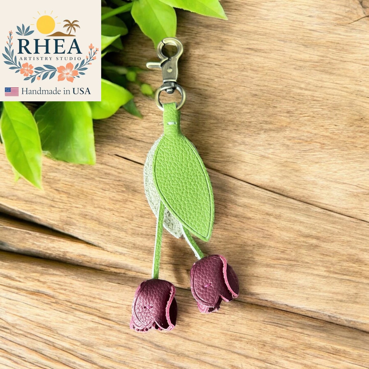 Green and purple leaf-shaped leather purse charm on a wooden surface with RHEA Artistry Studio branding.
