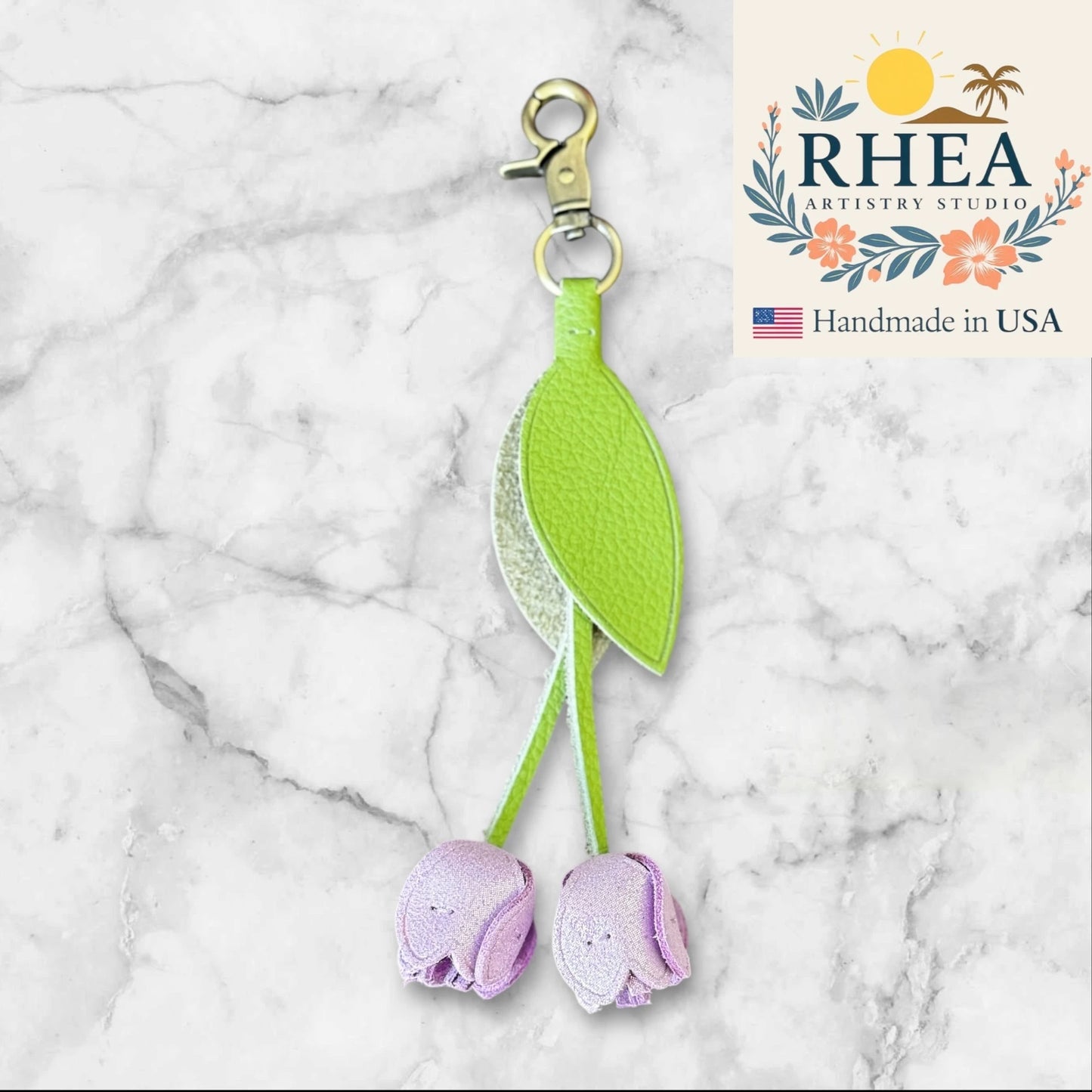 leather purse charm with green leaves and purple flowers on a marble background, featuring RHEA Artistry Studio logo.