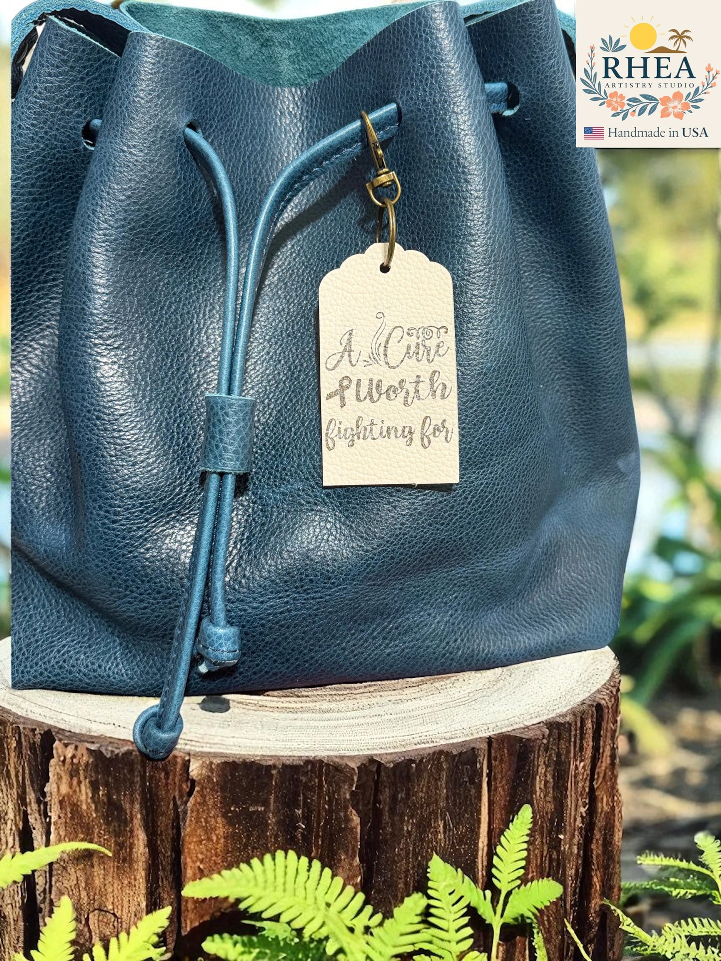 Blue leather bag with a tag on a wooden stump with plants in the background, featuring the RHEEA brand.