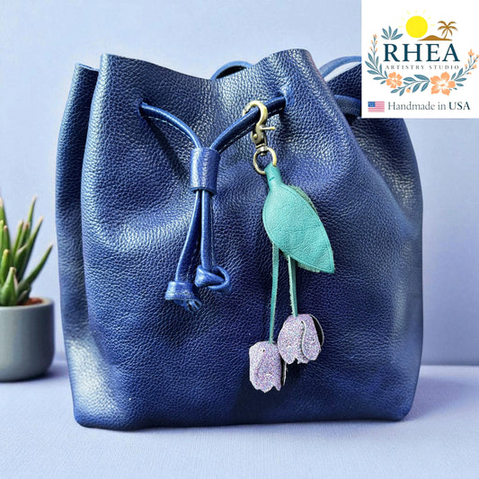 Blue leather bag with floral leather bag charm on a light blue background, featuring the RHEA brand.