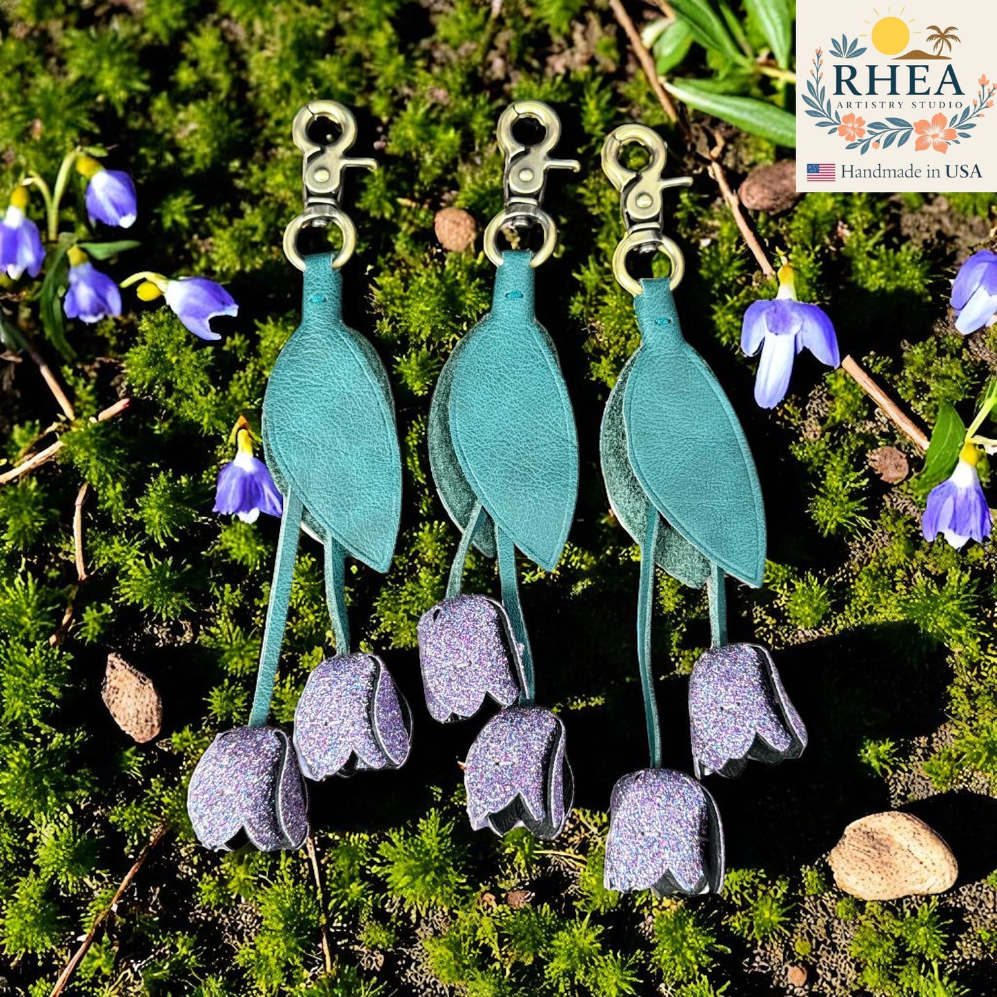 Teal and gray floral leather purse charms on a natural background with RHEA branding.